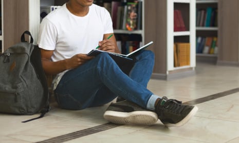Male student writing essay, sitting on floor at library