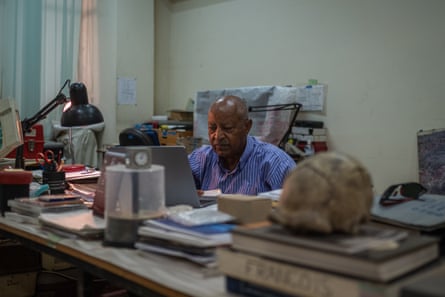 A man in a shirt sits at a desk at a computer. There are piles of books and papers on the desk, and a model of a skull in the foreground