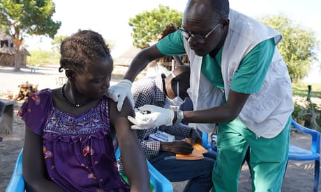 A health worker wearing green scrubs stands to inject the arm of a seated woman.