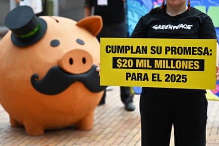 A person in a Greenpeace T-shirt holds a yellow sign with wording in Spanish while in the background there is a large ‘piggy bank’ with a top hat and moustache