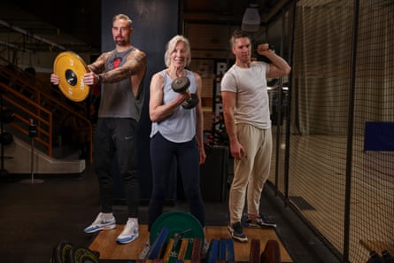 A tall woman with white hair holding dumbbells in a gym, with tall men on either side of her holding weights