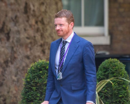 he wears a royal blue suit and dark blue tie with a lanyard round his neck; he is in his 40s and has short reddish hair and beard.
