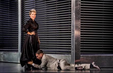 A singer in her mid-50s, with spiky blond hair, wearing a black ballgown, standing with one hand on hip looking smug, while a younger male singer in a grey suit prostrates himself at her feet, clutching one calf.