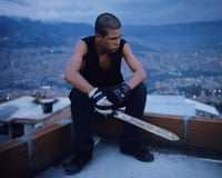 A young man sits on a rooftop ledge holding a machete, overlooking Barrio Triste at dusk