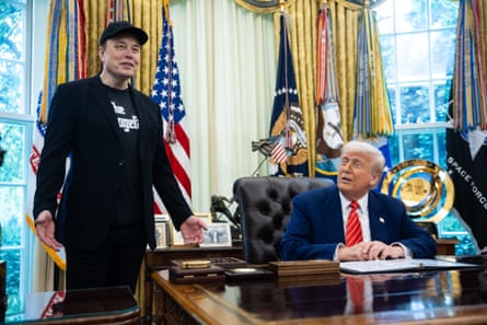 Musk standing, with Trump sitting at a desk looking up at him.