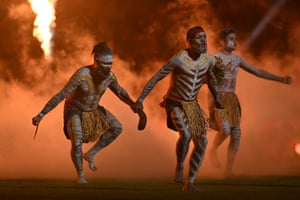 Indigenous dancers perform at a State of Origin game in Brisbane. Three dancers in cultural dress and paint are dancing while enveloped in orange smoke, with fire in the background