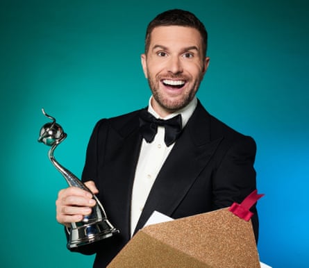 The presenter, in black bowtie and dinner jacket, holds an award and a glittery envelope