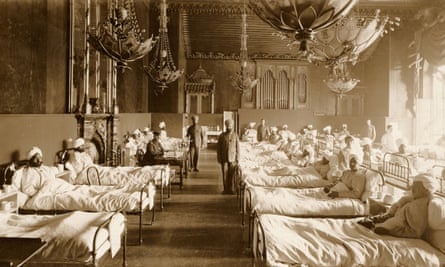 The image shows rows of beds containing wounded Indian soldiers. Two soldiers in turbans are standing halfway along the rows towards the centre of the image and a British soldier is seated behind a desk before the fireplace.