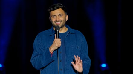 man in blue denim shirt speaks into microphone with hand raised on stage