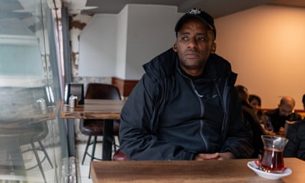 Siyabonga Twala sitting alone in cafe with a cup of coffee on table in front of him; he is wearing a black baseball cap and jacket and looking out of the window