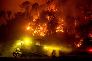 A bulldozer clears vegetation as a wildfire burns close by against a dark orange sky