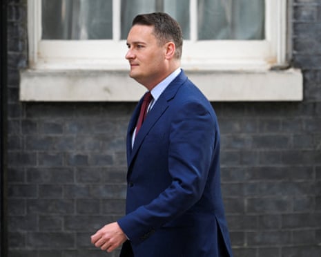 Streeting in a dark blue suit walks in profile past a brick building.