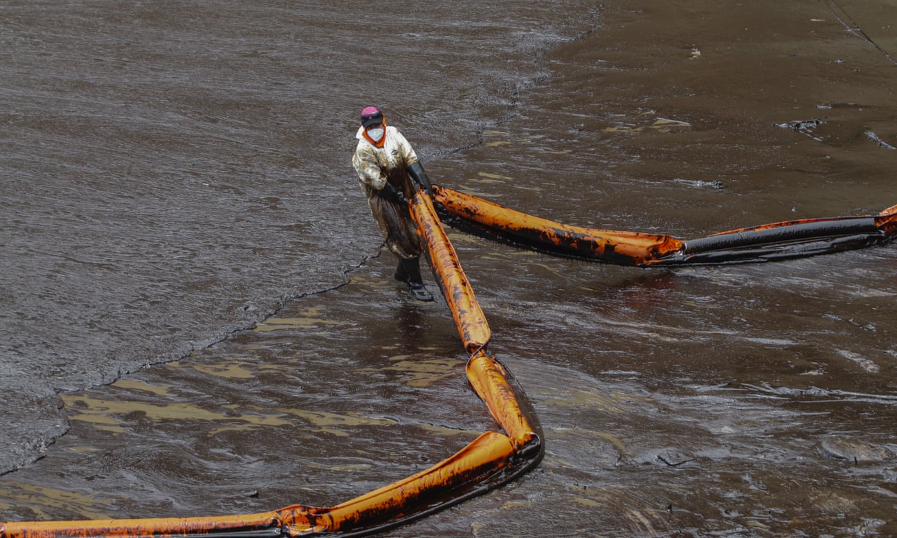 A worker on an oily beach drags a boom across slick-covered water