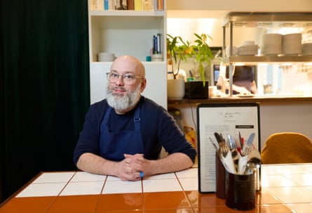 Man with a grey beard in a blue apron sat in a cafe