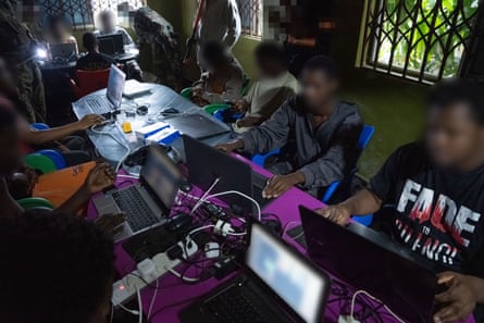 Suspected scammers sit with multiple laptops around tables at a raided scam centre