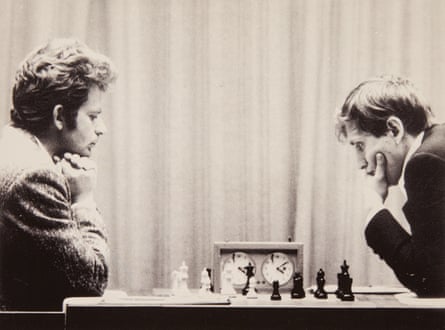 Spassky, on the left with chin resting on his hands, faces Fischer across the chess board. The American is holding his chin as his arm rests on the table. Spassky is playing with white, Fischer black