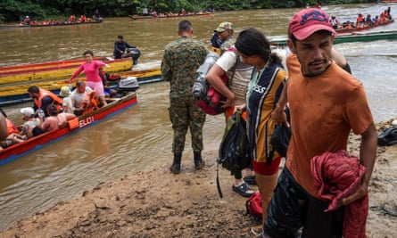 A group of long, skinny, brightly painted canoes disgorge passengers from a muddy river to a sandy bank. In the foreground is a brown-skinned man with a thin black mustache, wearing an orange T-shirt and pink baseball cap. He stands in a short line of other migrants.