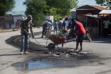 Men with a wheelbarrow a broom work amid water on an asphalt road.