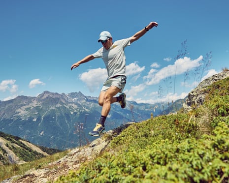John McAvoy leaps down a grassy mountainside with arms outstretched, peaks behind him