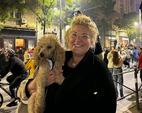 Lara smiles while holding her poodle on a busy night-time street in Belgrade