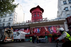 A cyclist in a hi-vis jacket and helmet looks up at the Moulin Rouge building, which is missing the M, O and U, and also the sails on its windmill. There is a barrier and construction tape outside it and what looks like the sails on the ground