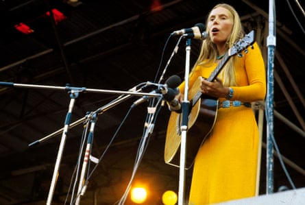 A woman with long blond hair playing guitar on stage wearing a sunshine yellow dress