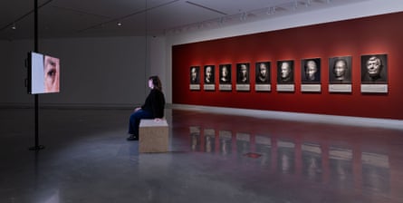 A visitor views a video artwork facing a red wall of nine portraits from Hoda Afshar’s Agonistes series