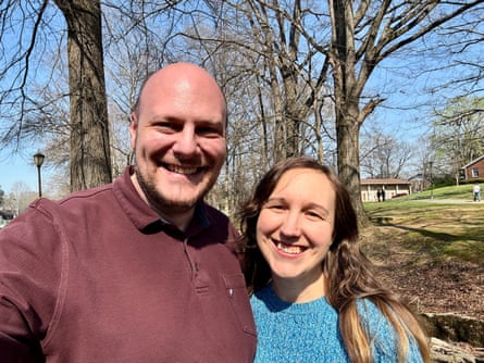 A man and woman standing close together, smiling, trees behind them