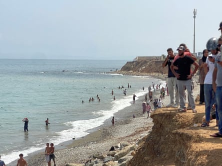 A group of people standing on a cliff with a beach and people swimming or walking in the sea below.