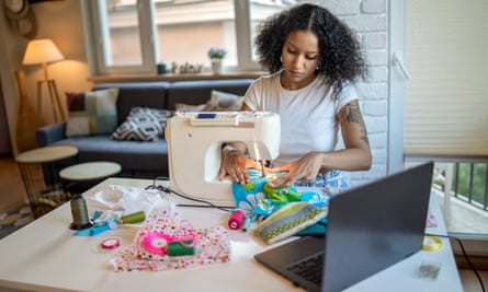 An African-American woman is using her sewing machine with her laptop close by on the table in front of her