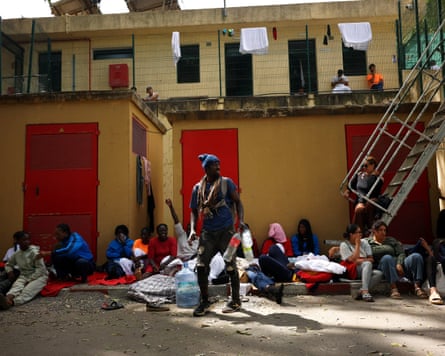 A man stands holding an empty Coke bottle near people sitting on the ground outside a building