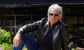 Jon Bon Jovi sits on a wall in a classic 80s rock star pose