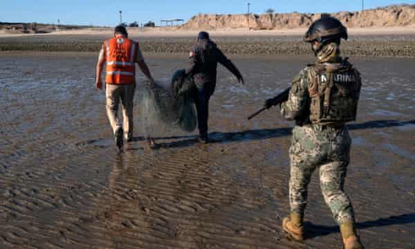 A man in camouflage with a gun walks behind two men carrying a large net on a beach