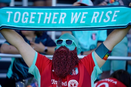 Teal Man, with his face painted in a teal colour and fan of Kansas City Current, holds a scarf aloft which says ‘Together we rise’ in the stands