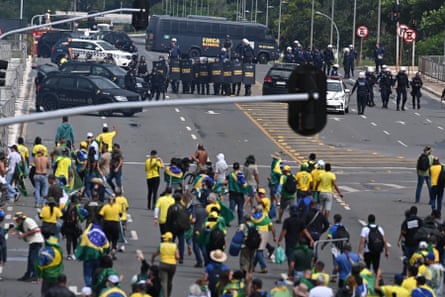 A large group of people, some draped in the Brazilian flag, stand at a distance from police on a road in Brasília