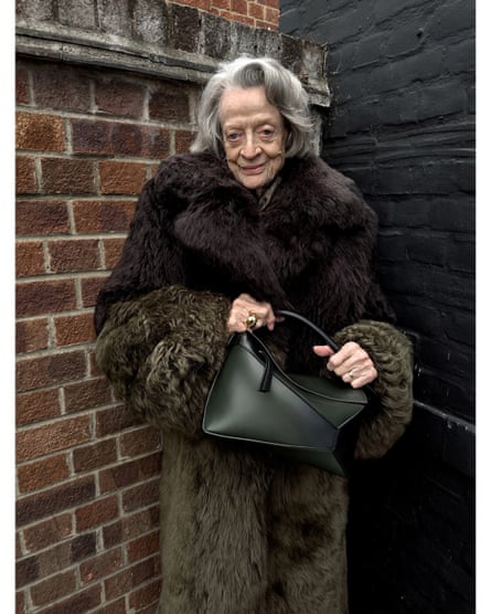 Maggie Smith in a large fur coat holding a black bag in front of a brick wall