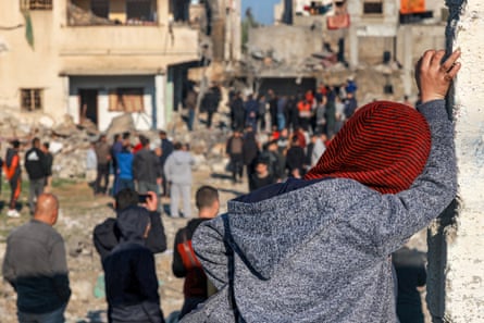 A woman leans against a wall watching in the distance a crowd of people mingling among the wreckage of a destroyed building.