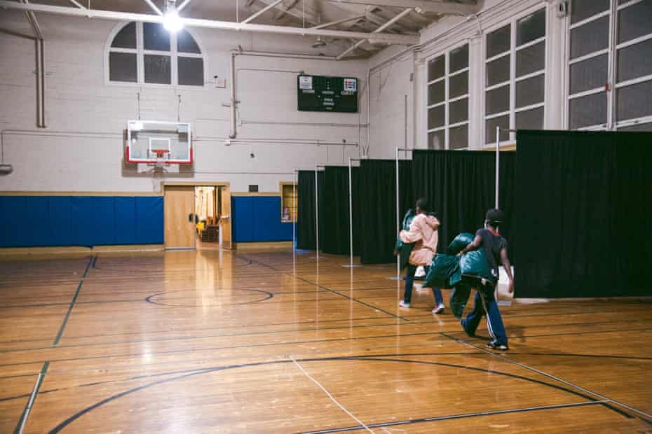 Two children holding sleeping bags walk through a gymnasium toward a corner where partitioned cubicles are set up.