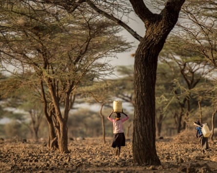 two girls walk away from a water source; one carries a large white container on her head and the other on her back. They walk through trees and bushes over rocky ground.
