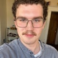 A person with short curly brown hair and a mustache who is wearing glasses poses for a selfie