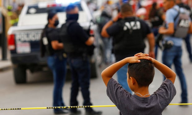 A small boy with his hands on his head stands behind yellow police tape, looking at the police activity in a parking lot.