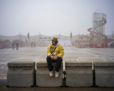 A young man sits on concrete blocks in front of a helter-skelter and an illuminated Blackpool sign on a misty day.