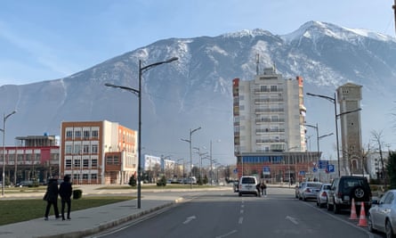 Photograph of an empty street with two people walkign along and two or three buildings against a backdrop of high mountains