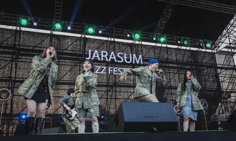 Four members of Leenalchi on stage, three women and a man, singing or rapping into their microphones, all wearing camouflage-style jackets over their various outfits