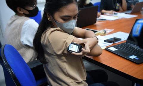 A young woman sitting with others at a large table holds an electronic device against her upper arm