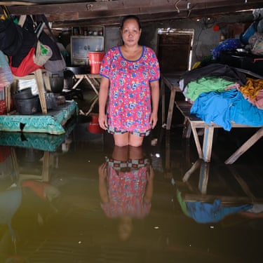 A woman in a pink dress stands in a room with flood water thigh-deep and things piled up on table tops