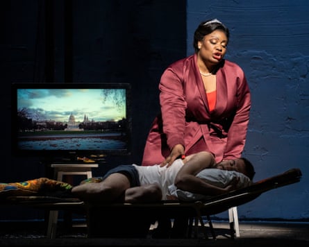 A opera singer wearing a tiara and a mauve silk dressing gown singers of a finger in underwear and white vest with a TV screen in background showing the US Capitol