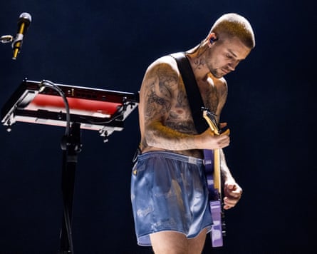 A shirtless Justin Bieber, wearing bluish shorts, strums an electrical guitar connected stage.
