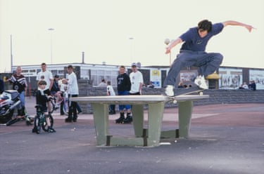 A person skateboarding on the corner of a table outdoors, with people looking on