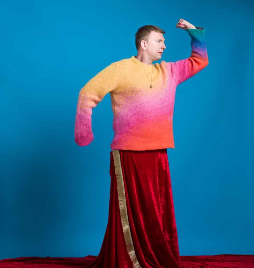 Joe Lycett in a superhero pose, wearing a multicoloured jumper and a red velvet skirt